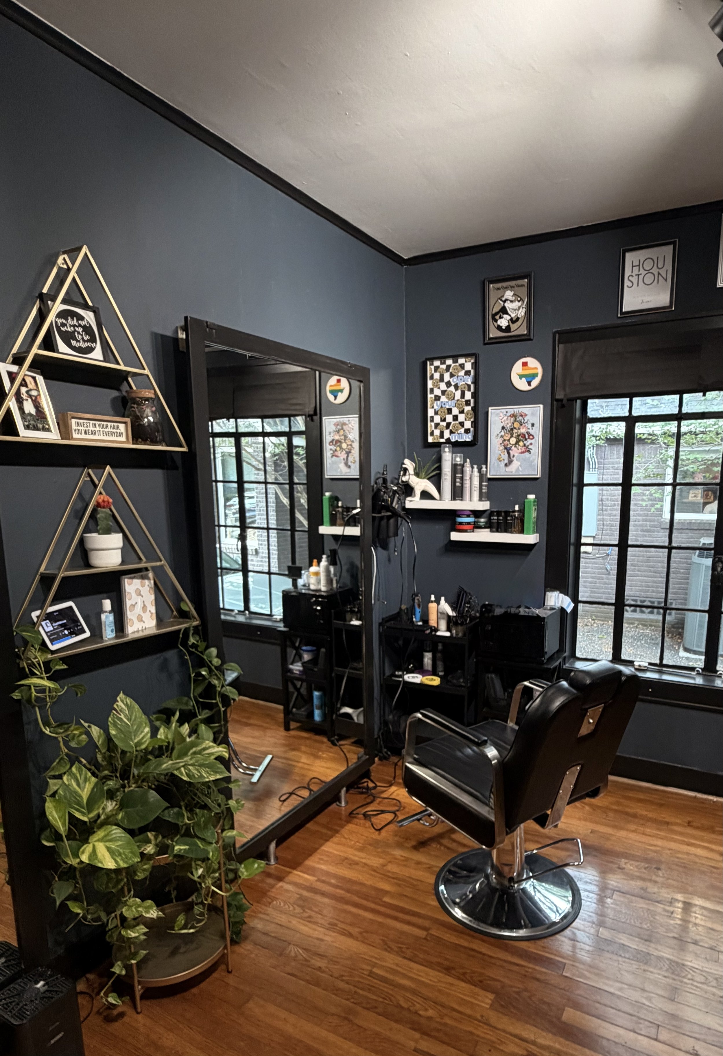 A hair salon with dark blue walls, a large mirror, and black styling chairs. There are framed pictures and artwork on the walls, a shelf with hair products, and a window with blackout blinds. Plants are visible, and the floor is hardwood.