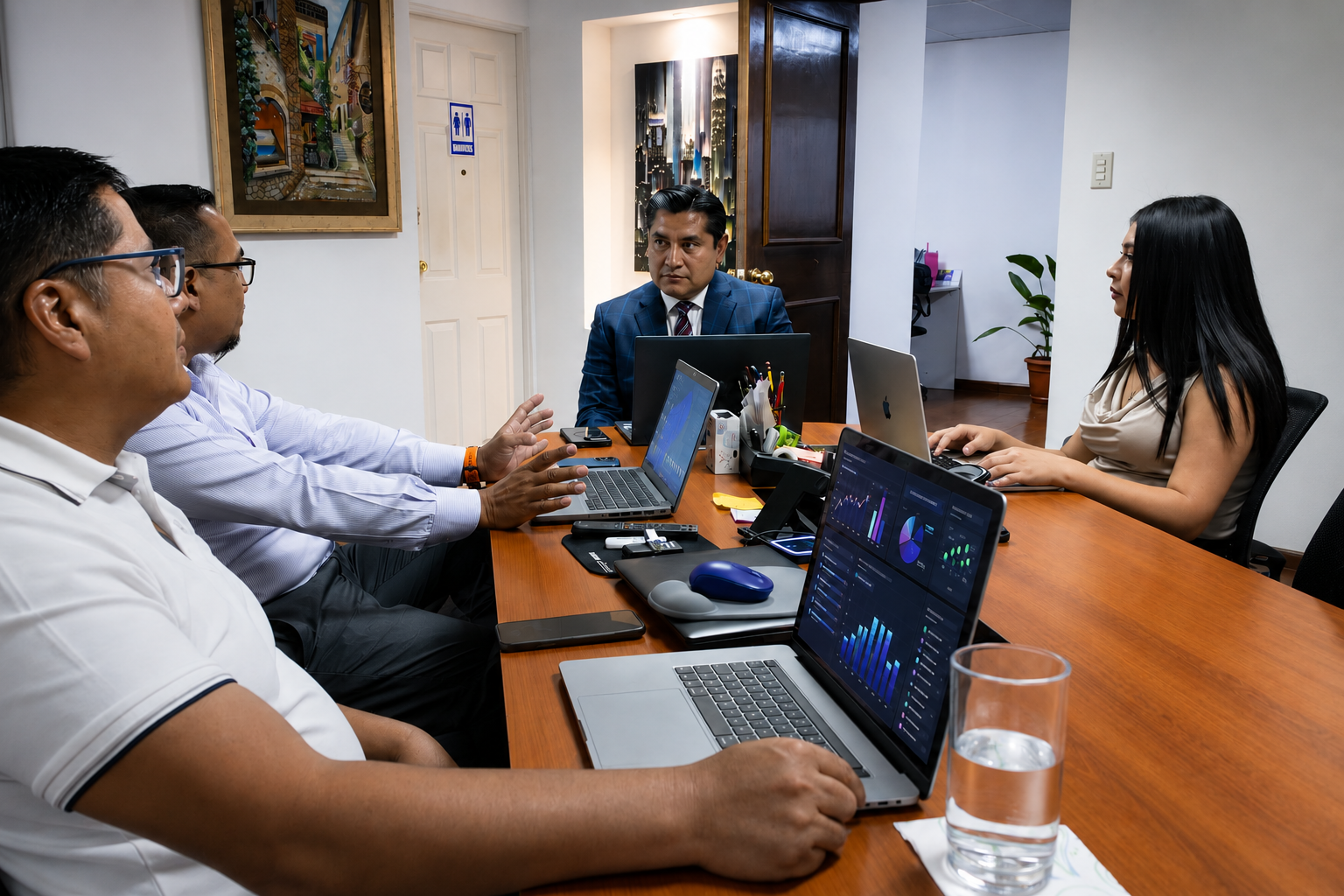 Reunión de cinco personas en una sala de oficina, sentadas alrededor de una mesa con laptops, gráficos y documentos, discutiendo en un ambiente formal.
