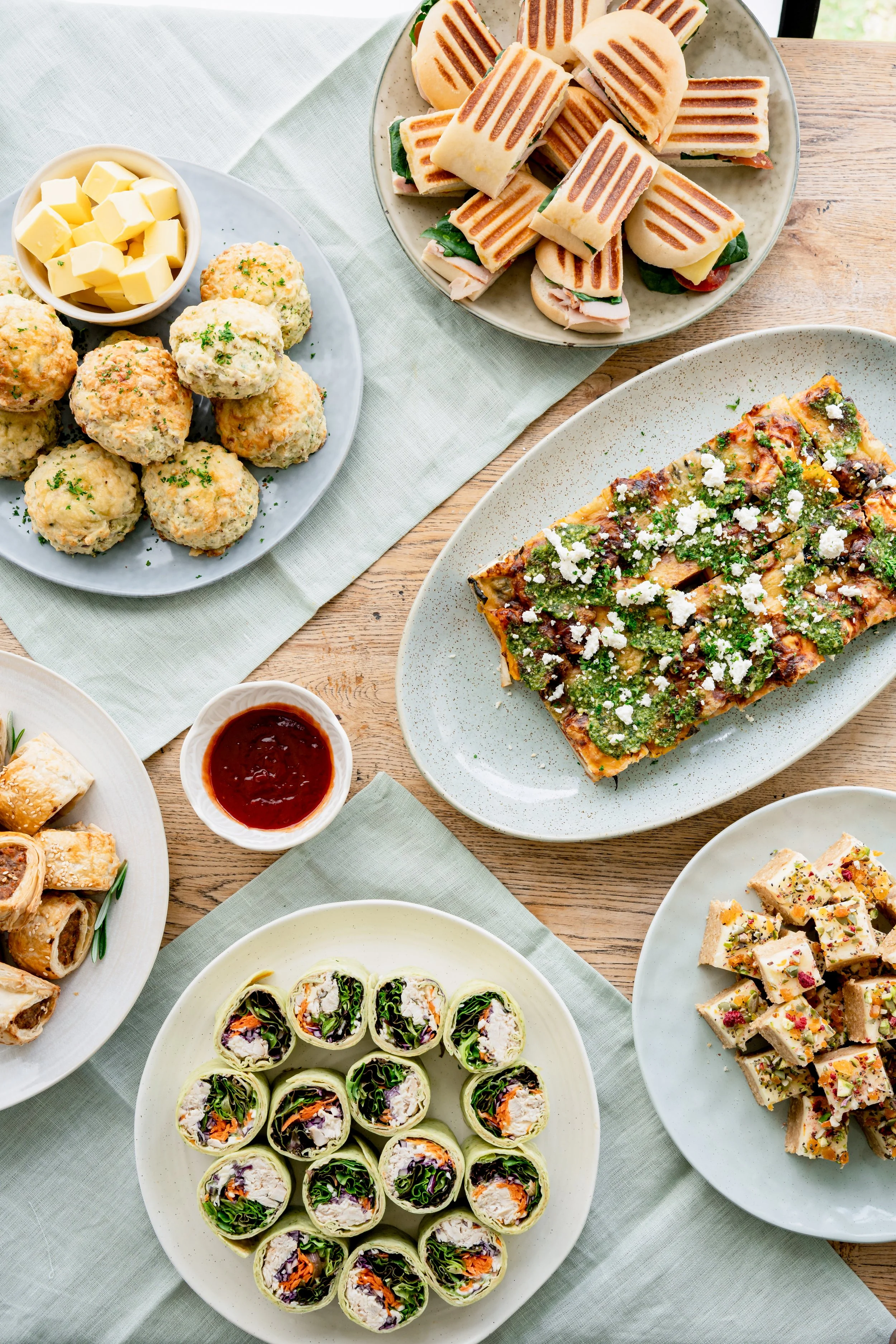Assorted dishes including a plate of sushi rolls, a dish of lemon pepper chicken, grilled sandwiches, and a plate of veggie wraps, with a bowl of dipping sauce on a wooden table with a green cloth napkin.