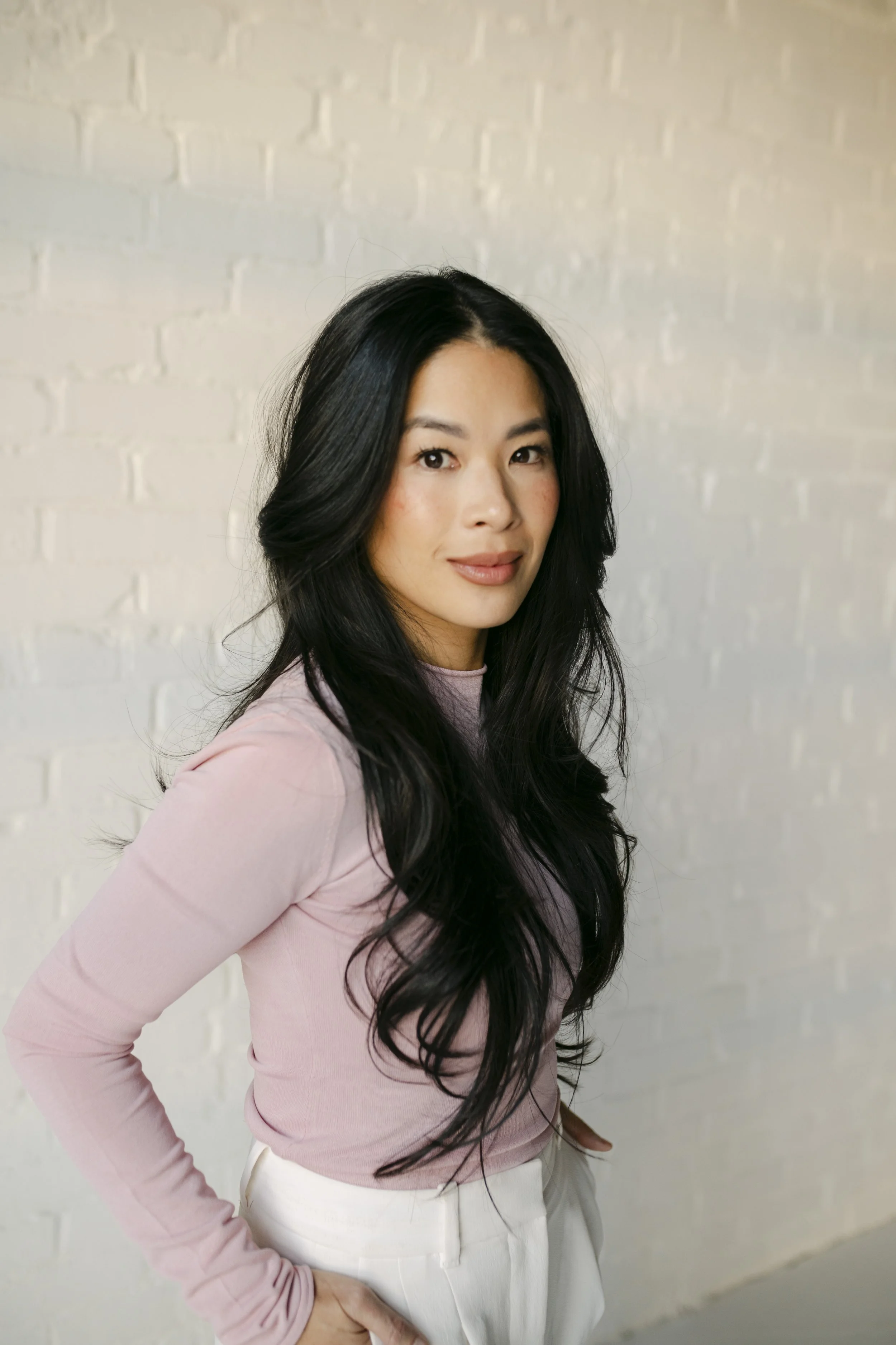 A woman with long black hair wearing a light pink long-sleeve shirt and white pants standing against a white brick wall.