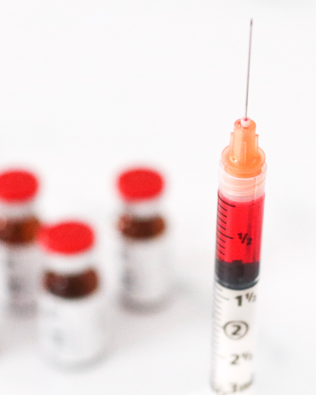 A medical syringe filled with red liquid, with a thin needle attached, next to several small vials capped with red lids.