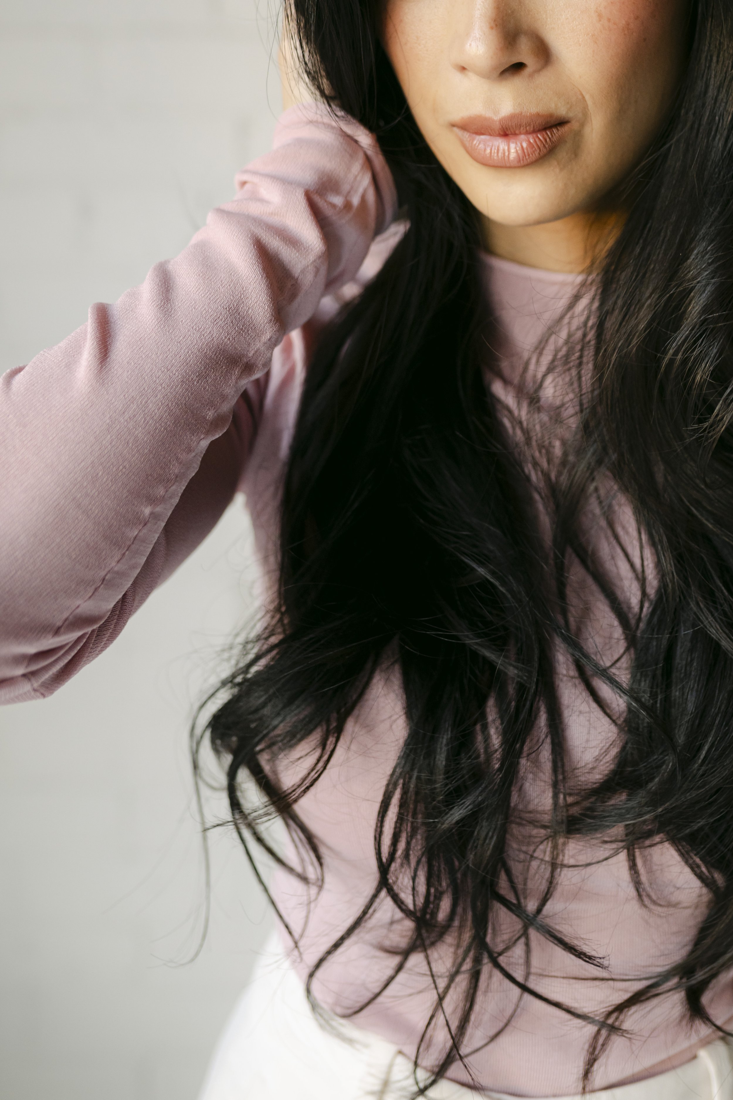 Close-up of a woman with long dark hair wearing a pink top, with part of her face showing, against a plain white background.