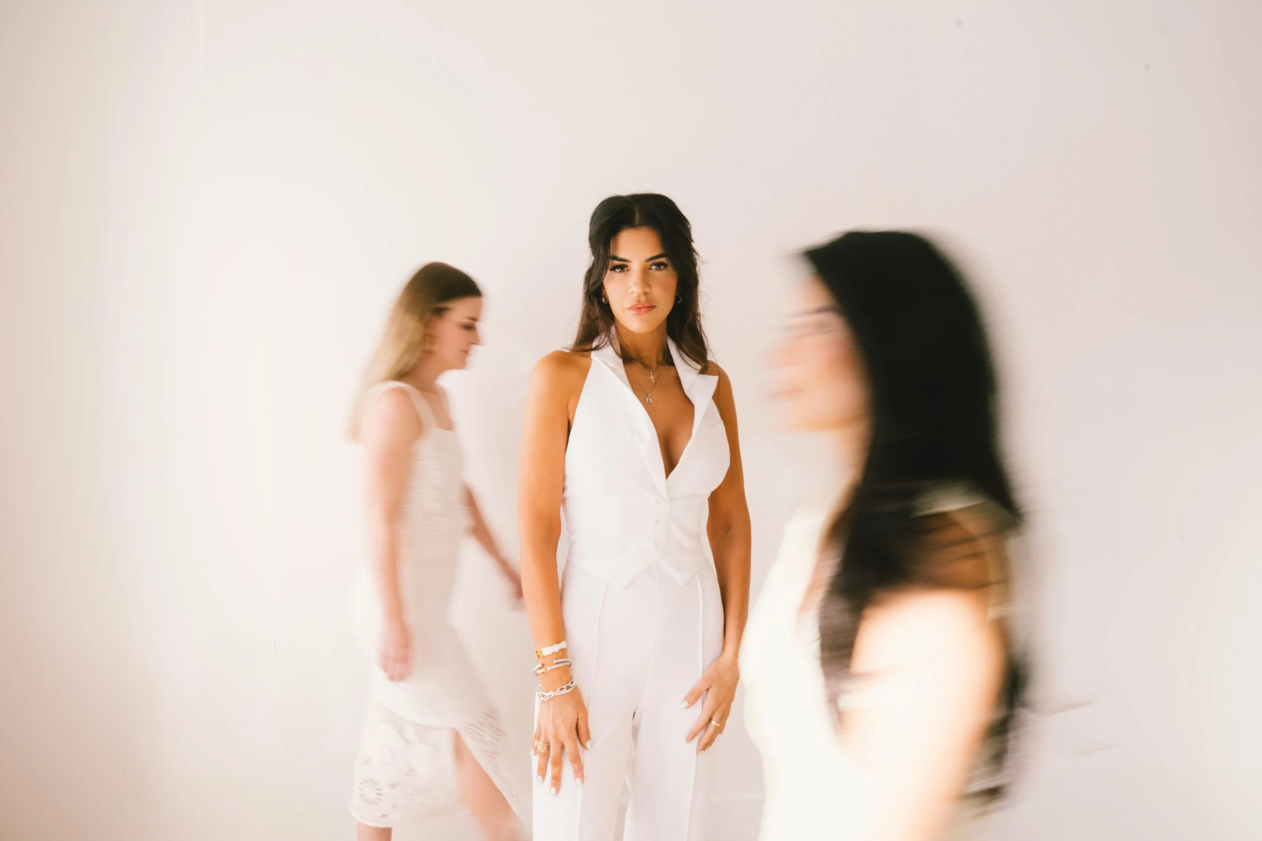 Rebecca Williford, owner of The Rae Clinic in Evans, Georgia, with long dark hair wearing a white sleeveless dress stands against a plain white wall, surrounded by blurred figures of other women.
