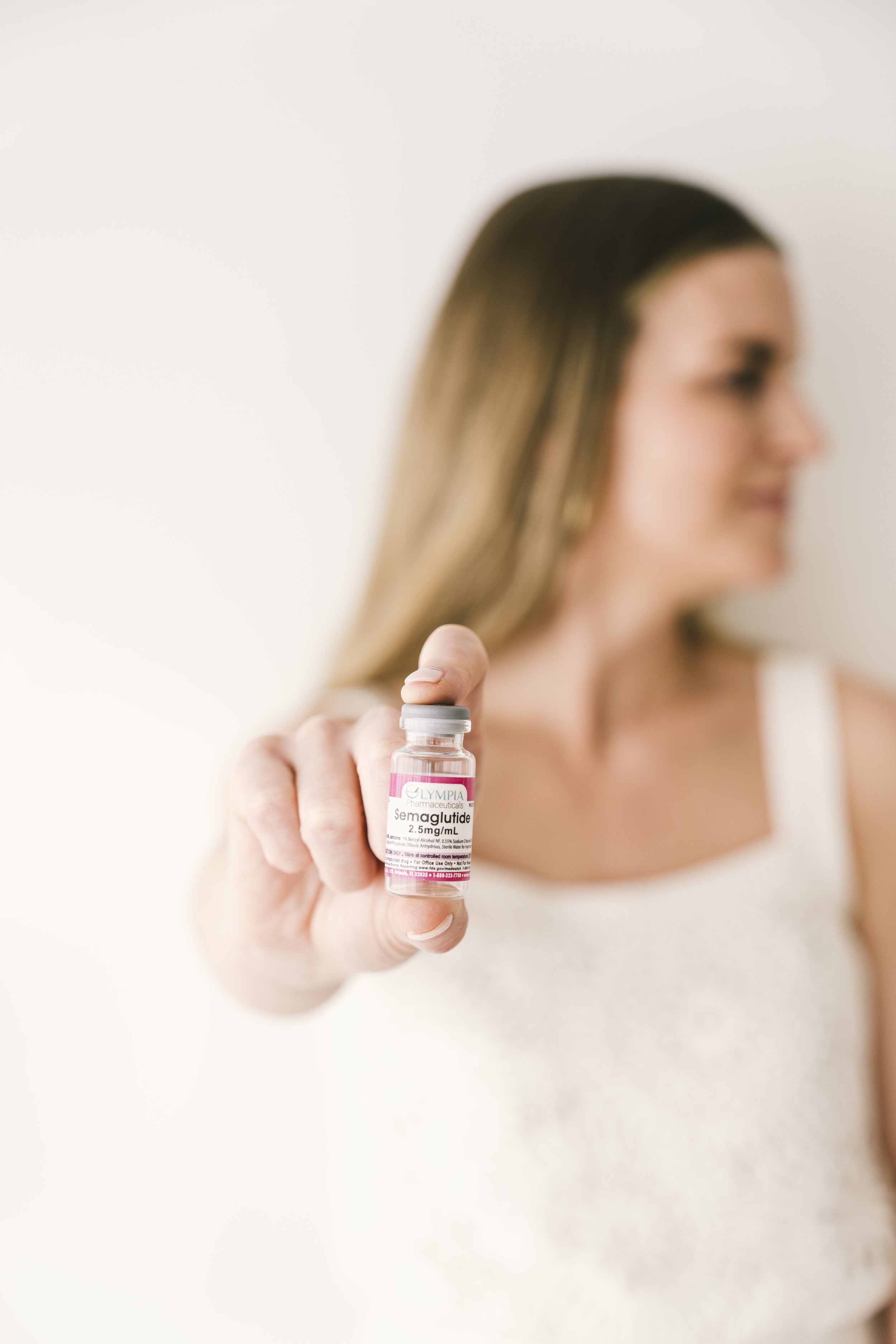 A woman holds a small vial labeled 'Semaglutide 2.5mg/mL' in front of her face, slightly out of focus.