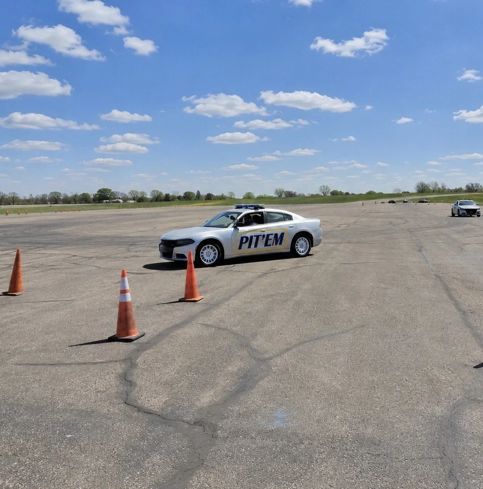 A police car marked 'PIT' in a large open parking lot with orange cones set up around it, under a partly cloudy blue sky.