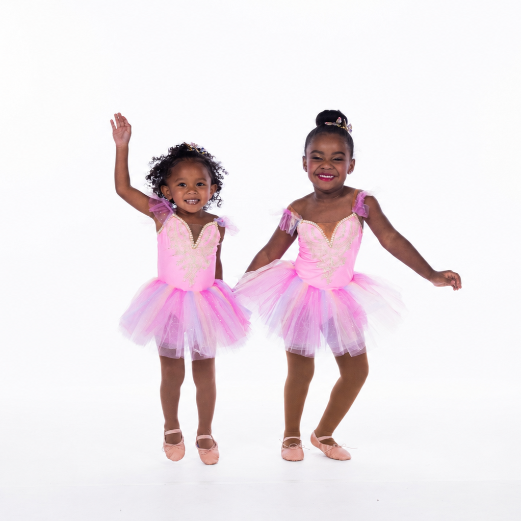 Two young girls in pink ballet costumes and tutus smiling and dancing against a white background.