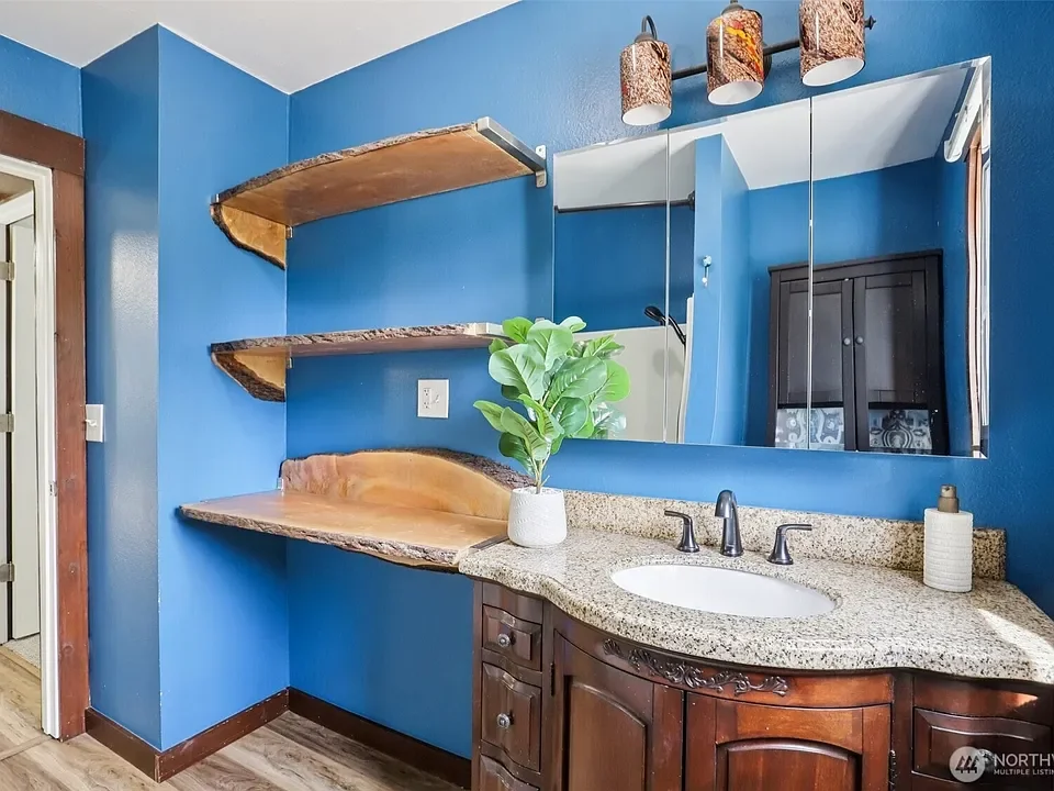 A bathroom with bright blue walls, a granite countertop with a white oval sink, a large mirror, dark wooden cabinetry, and open wooden shelves with live-edge wood finishes. A small potted plant is on the counter, and three pendant light fixtures are 