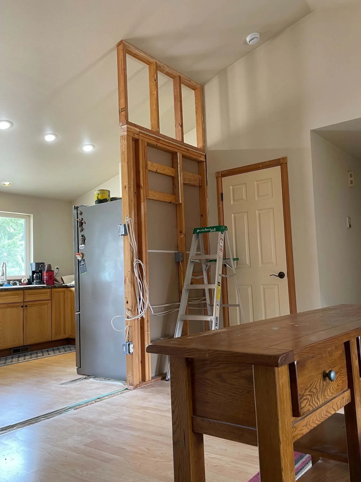 Kitchen under renovation with exposed wall framing, wiring, refrigerator, ladder, and wooden island