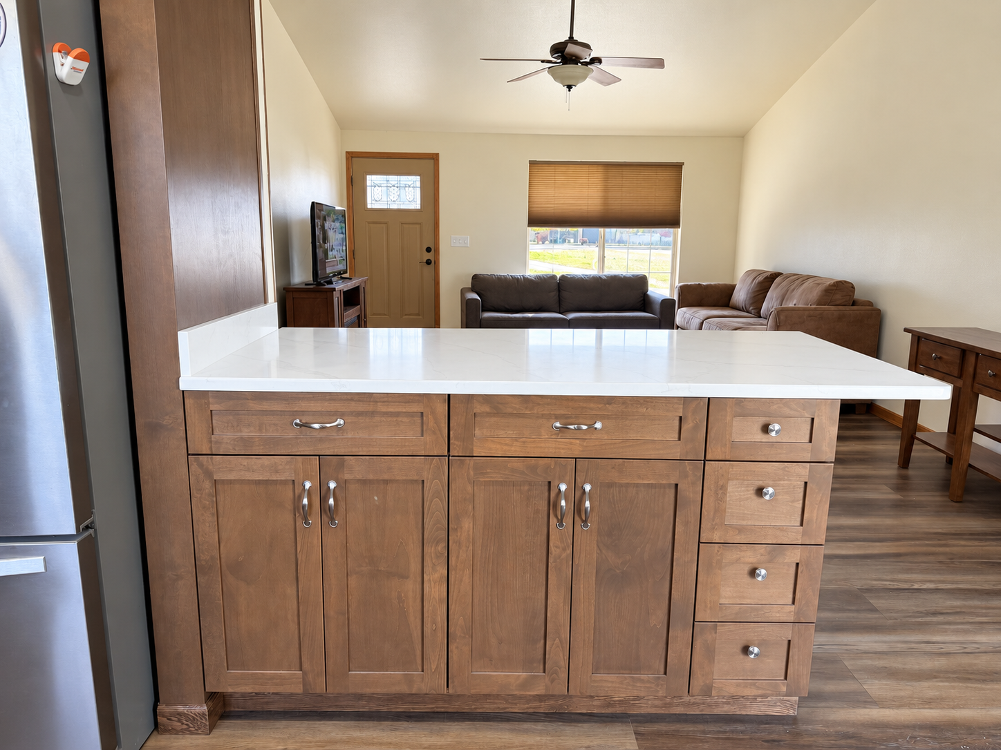 Kitchen island with white quartz countertop and wood cabinets overlooking a living room with sofas, TV, ceiling fan, and window.