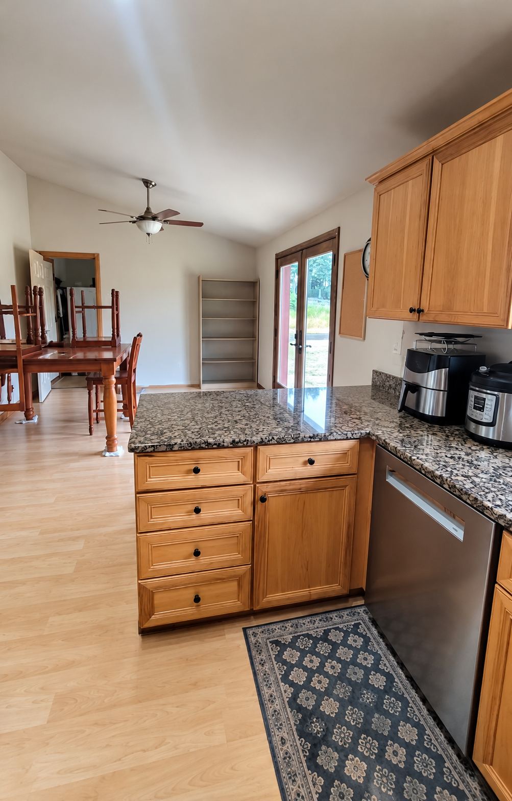Bright kitchen with honey-colored wood cabinets, granite island, stainless steel dishwasher and small appliances, adjoining dining table, ceiling fan, and glass exterior door.