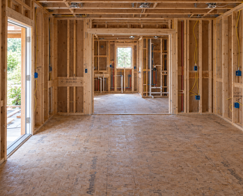 Interior view of a house under construction, showing exposed wooden framing, electrical wiring, and doorways leading to other rooms.