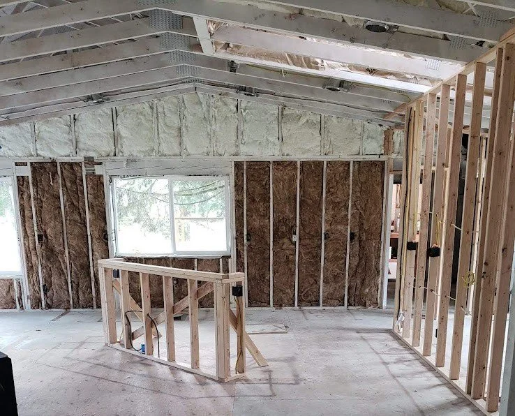 Interior of a house under construction with exposed wall studs, ceiling joists, and insulation, and a window in the background.