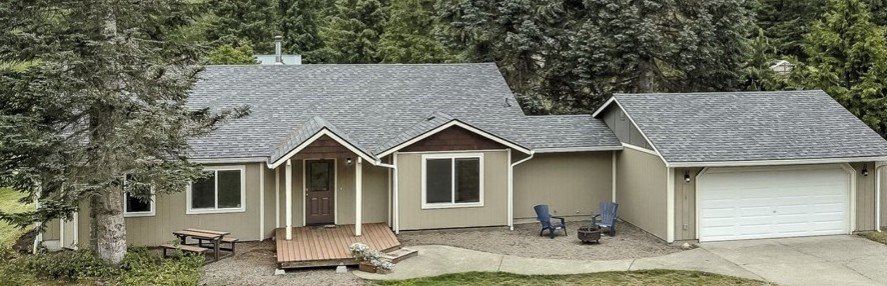 A single-story house with beige siding, a front porch, a two-car garage, and surrounding trees.