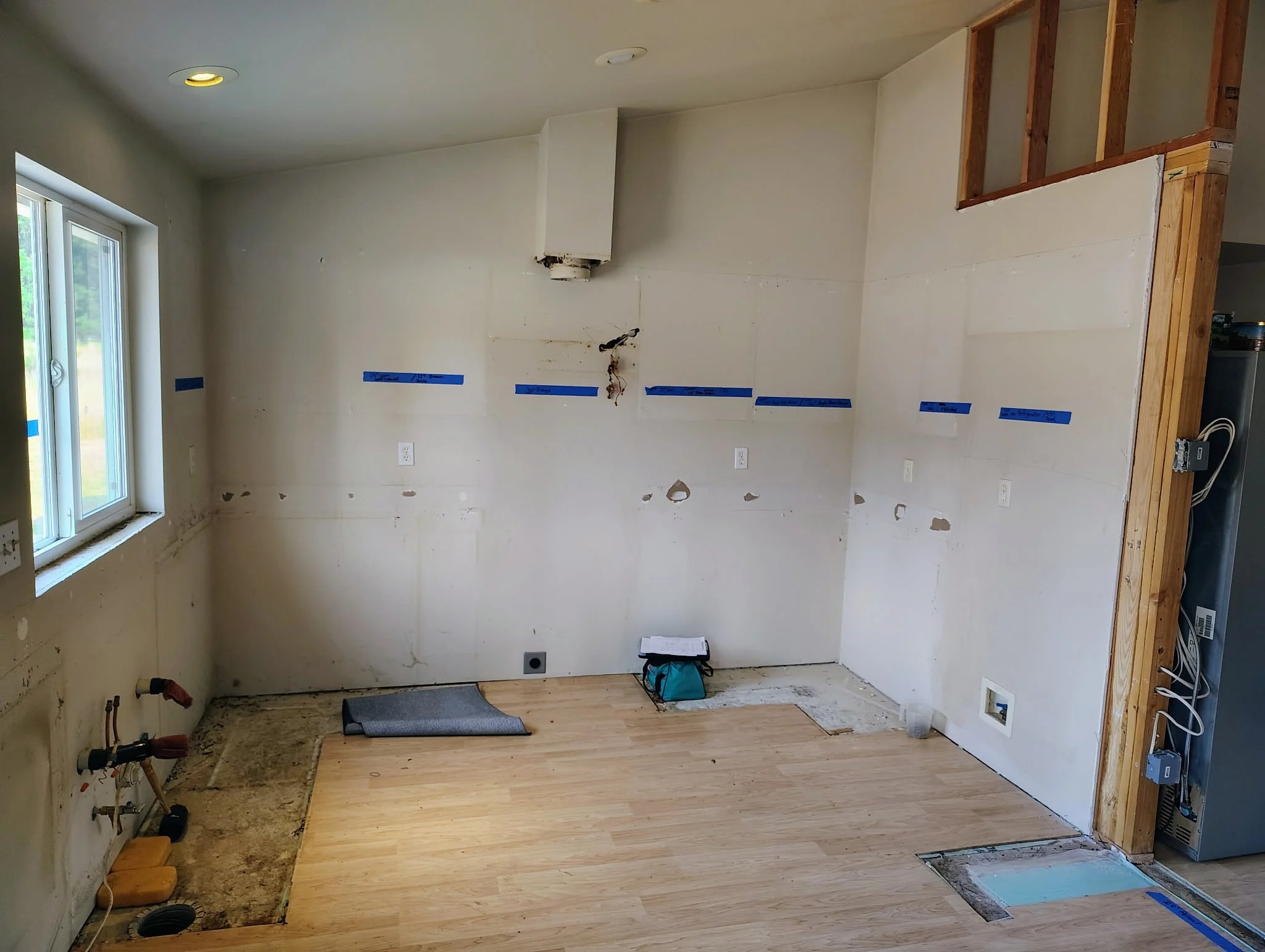 Partially renovated kitchen with exposed plumbing and electrical wiring, unfinished walls, open framing, and newly installed wood flooring.