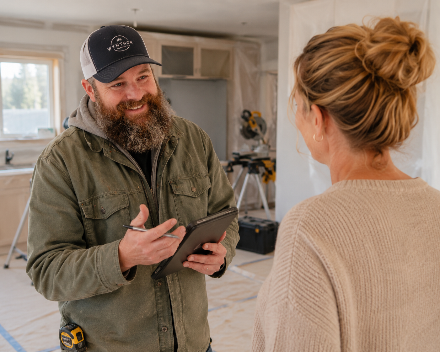 A smiling man with a beard and wearing a cap and work jacket talking to a woman with curly hair in a beige sweater in a room under construction.