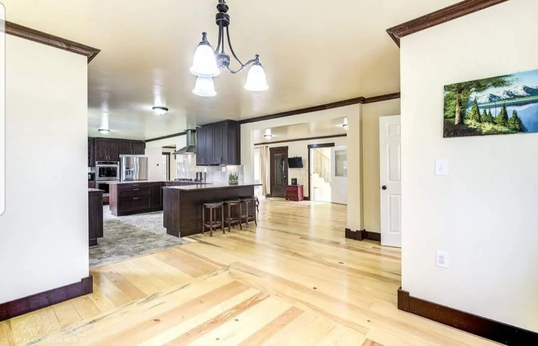 Open-concept kitchen and living area with light-colored hardwood floors, dark wood cabinets, stainless steel appliances, and a hanging chandelier with multiple lights.