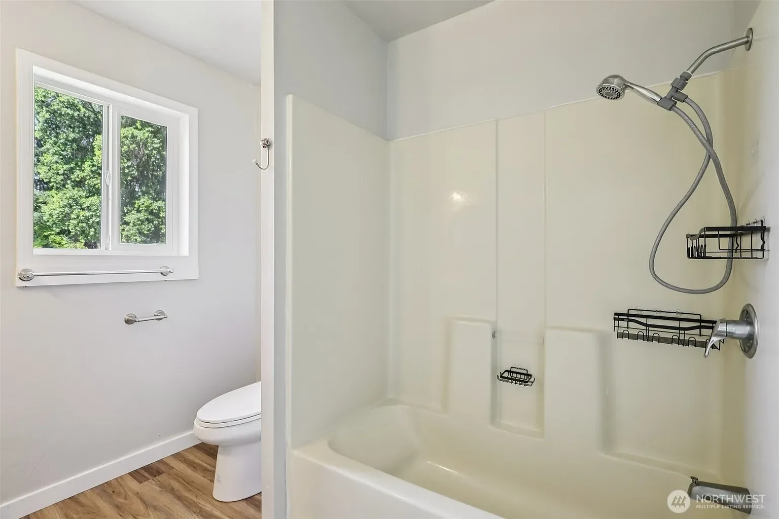 Bathroom with white walls, wooden floor, window, toilet, and shower with handheld showerhead and storage caddies.