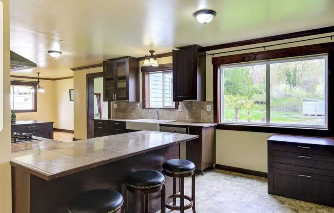 Kitchen with dark wooden cabinets, a large window showing outdoor greenery, a tiled countertop, and bar stools.