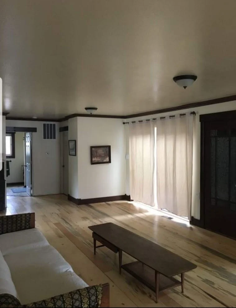 Empty living room with a wooden floor, a couch on the left, a coffee table in the center, and curtains covering a sliding glass door on the right.