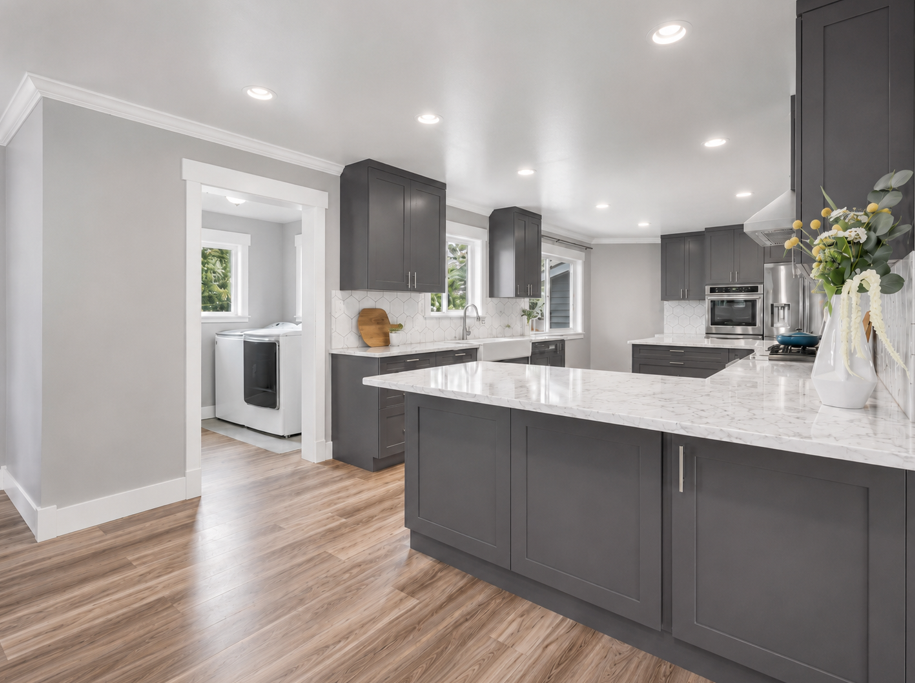 Modern kitchen with gray cabinets, white marble countertops, and hardwood floors. There is a small laundry area with a washing machine visible through an open doorway. The kitchen features a vase with flowers on the counter and multiple recessed ceil