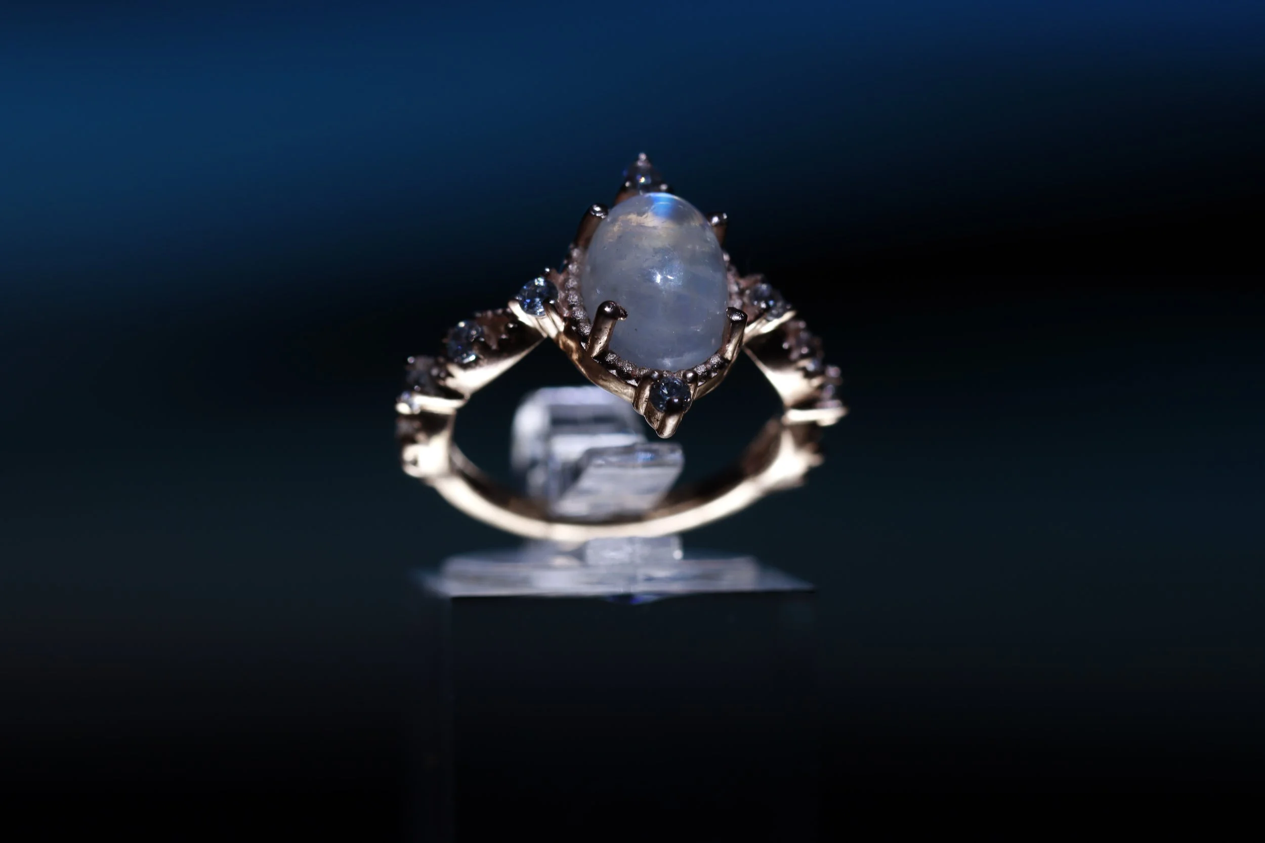 A ring with a large oval-shaped gemstone set in a detailed metal band, displayed on a transparent stand against a dark background.
