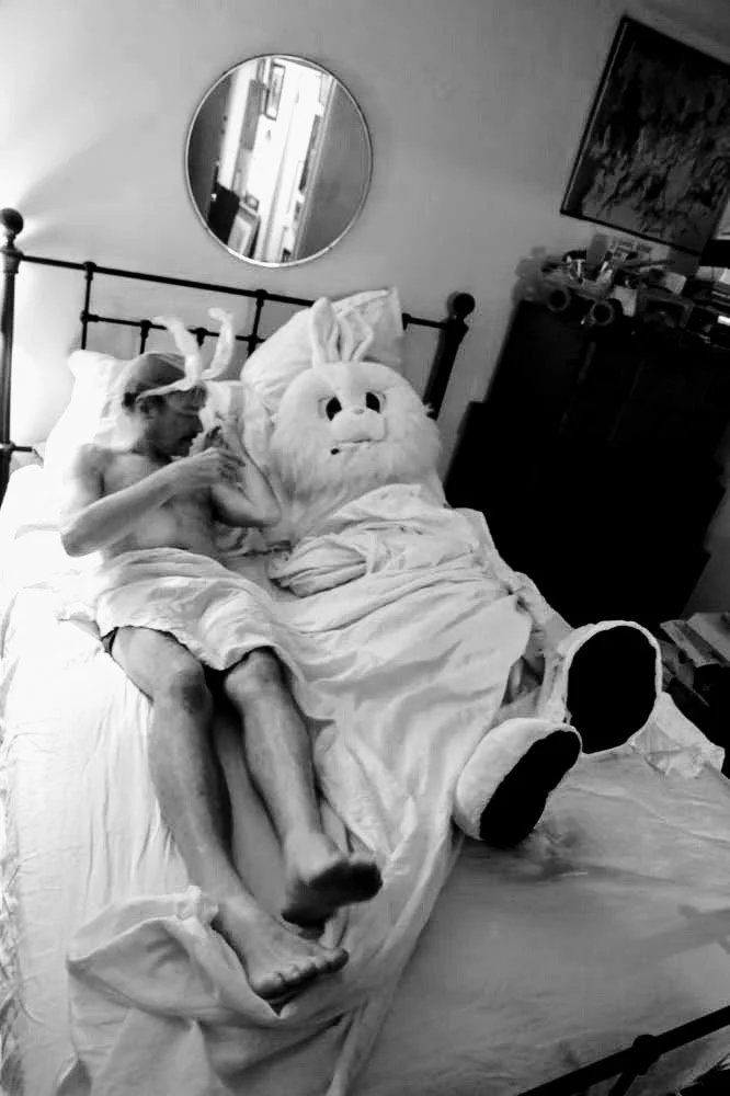A shirtless young boy with a bandage wrapped around his head, lying in bed next to a large plush bunny wearing a Santa hat. The boy appears to be giving a high-five to the plush bunny.