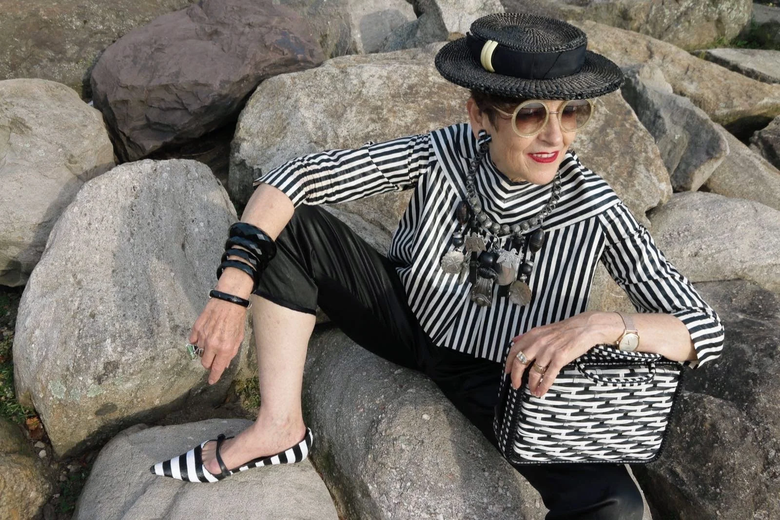 Woman wearing black and white striped top, black pants, black and white striped shoes, large hat, sunglasses, and jewelry, sitting on rocks with a striped bag.