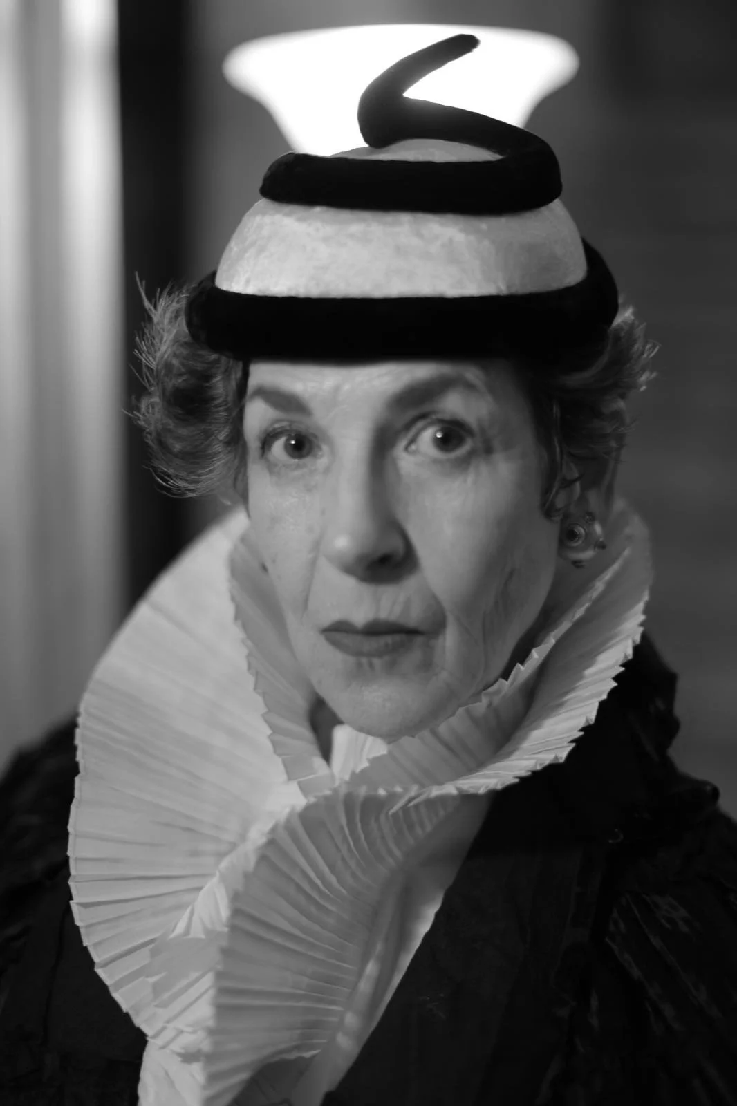 A black and white photo of a woman wearing a hat with a small brim, decorated with a large spiral-shaped ribbon, and a ruffled collar made of pleated fabric.