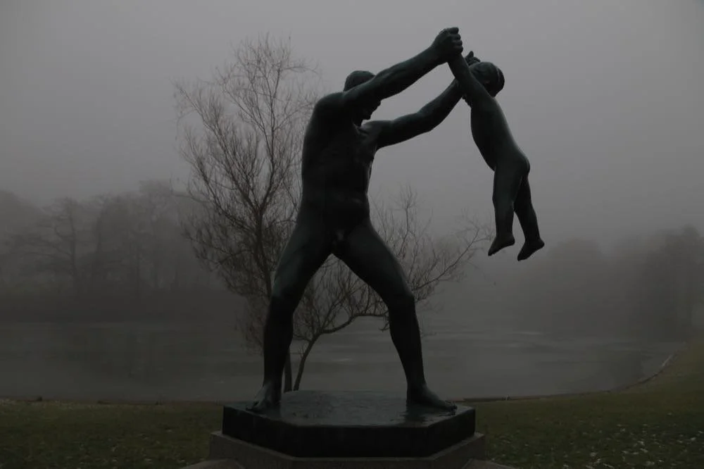 A statue of a man holding and lifting a child in the air, set against a foggy background with trees and a body of water.