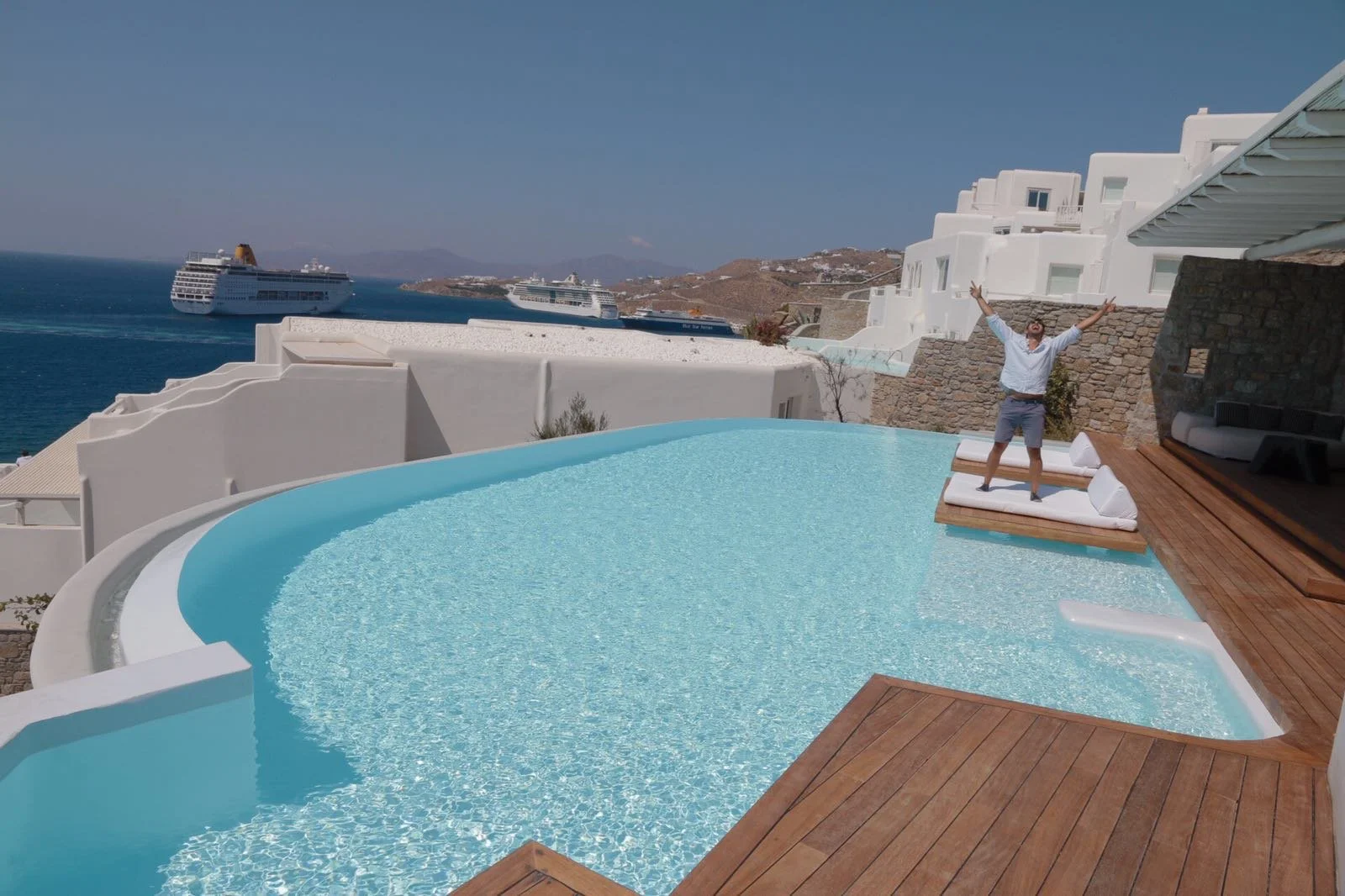 A man standing on a wooden lounge chair at the edge of an infinity pool with a view of the sea and cruise ships in the background on a sunny day.