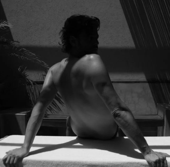 Black and white photo of a shirtless man sitting on the edge of a surface, viewed from the back, with a mural of palm trees in the background.