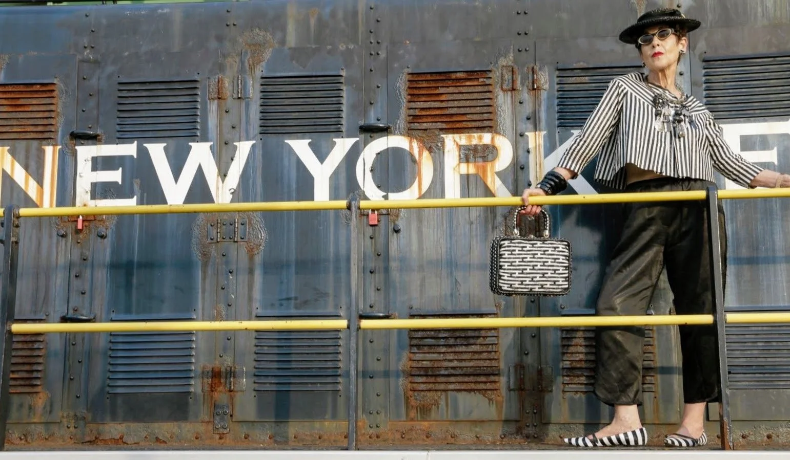 Woman dressed in black and white striped clothing, wearing sunglasses and a black hat, standing on a metal platform in front of a rusted train with 'New York' written on it, holding a patterned handbag and wearing striped shoes.