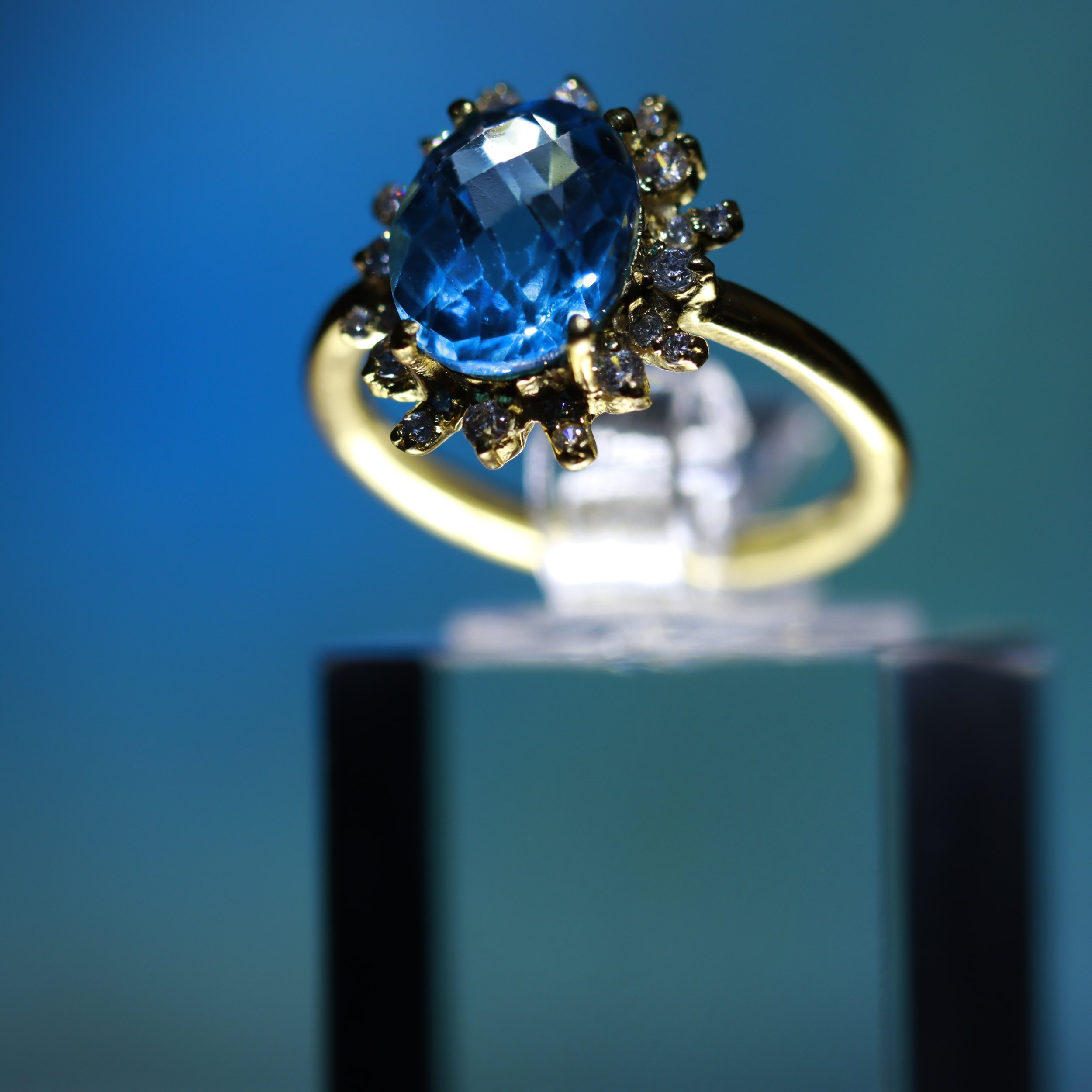 Close-up photo of a gold ring with a large oval blue gemstone and small clear stones around it, against a blurred green background.
