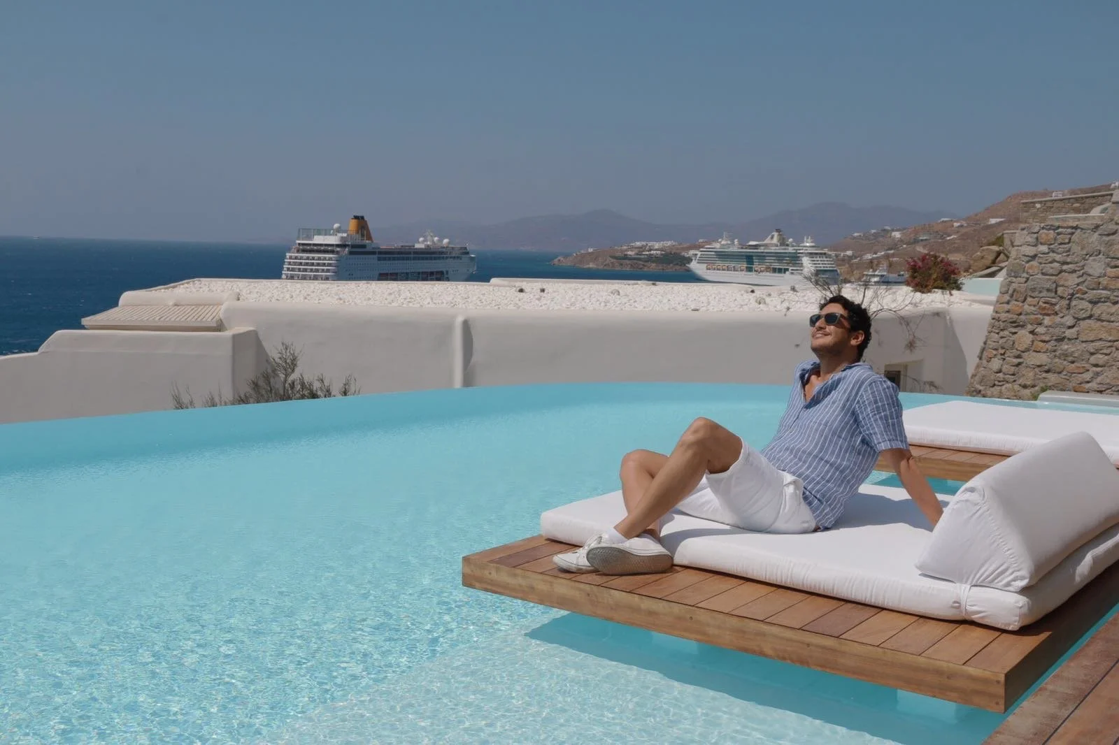 A man relaxing on a lounge chair by a swimming pool with a view of the ocean and cruise ships in the background.