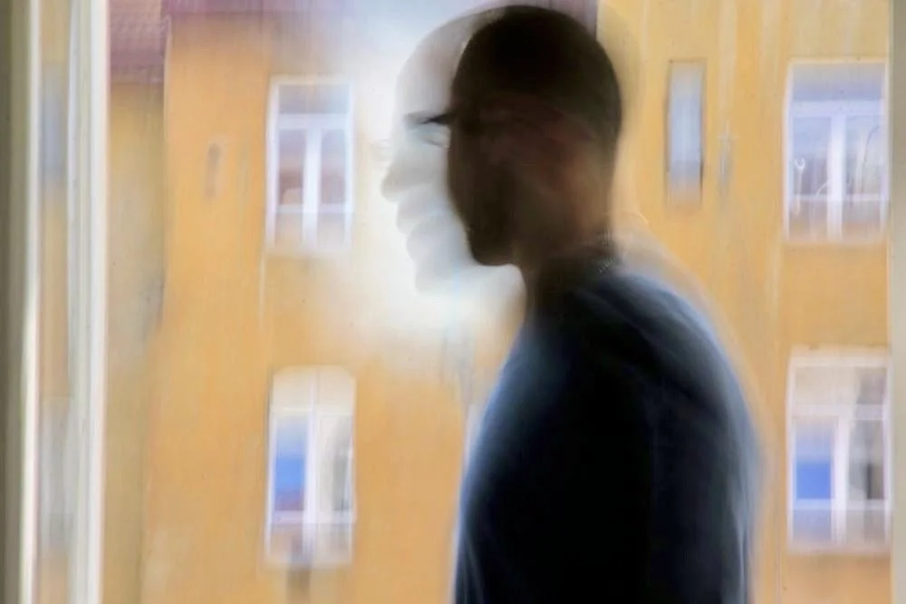 A blurry side view of a man with glasses and dark hair, standing indoors near a window with a yellow building outside.