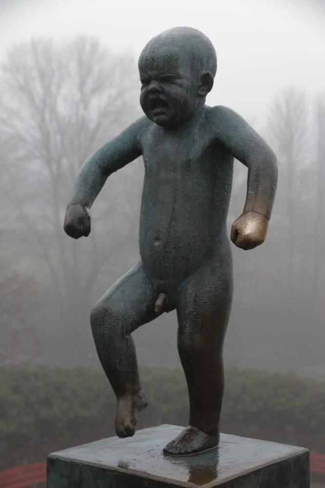 A sculpture of a young child, with a serious or angry expression, standing with one leg raised as if in motion, made of dark metal with some gold accents on the fists, set outdoors with leafless trees in the background.