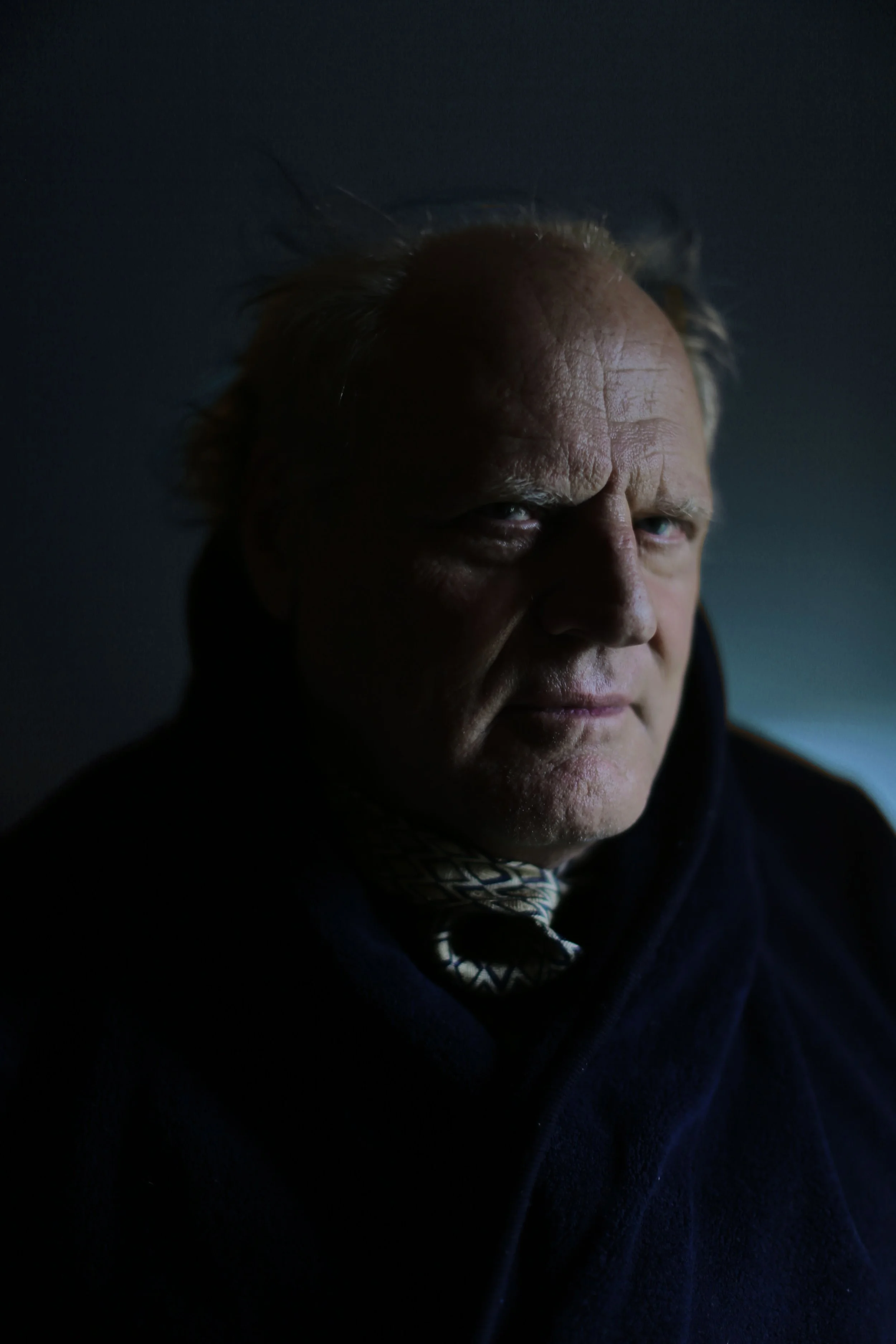 Close-up of a middle-aged man with a stern expression, light brown hair, and a focus on his face, wearing a dark jacket and sitting in a dimly lit environment.