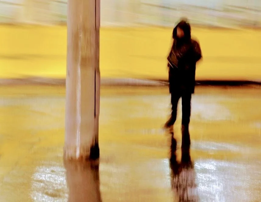 Blurred image of a woman standing on a wet street at night with reflections, near a large pole.