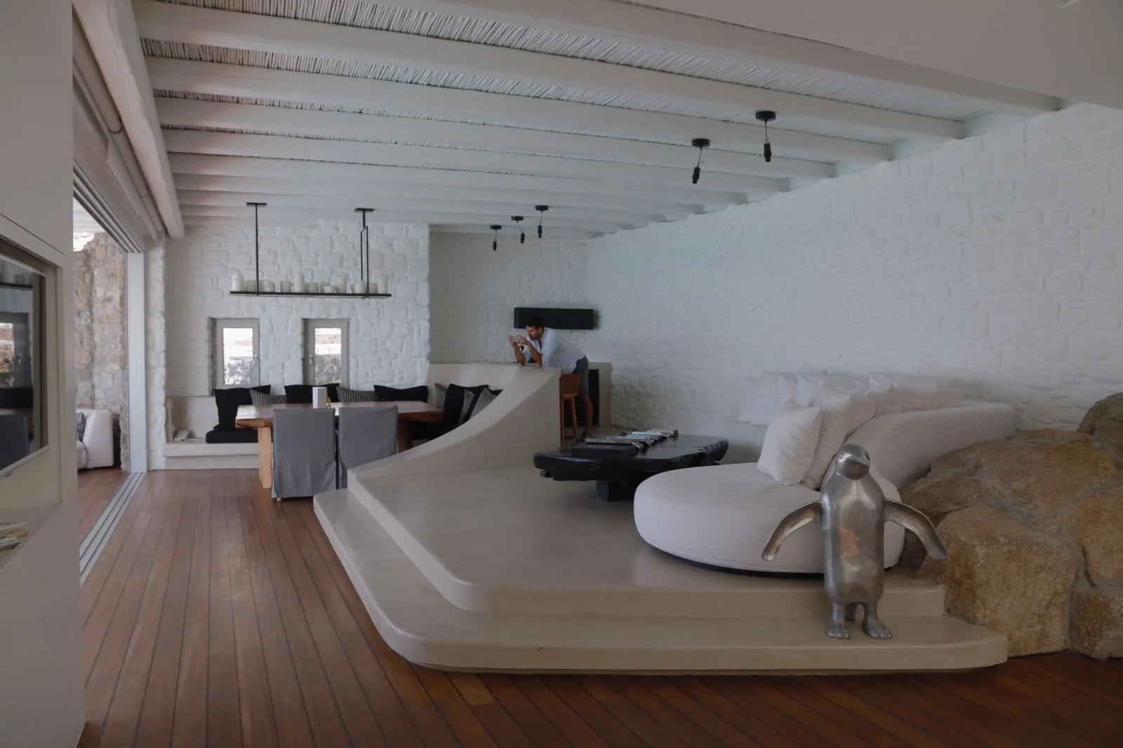 Modern living room with white brick walls and wooden ceiling beams, featuring a large white curved sofa, decorative sculptures, a dining area with black chairs, and a man standing near a bar area.