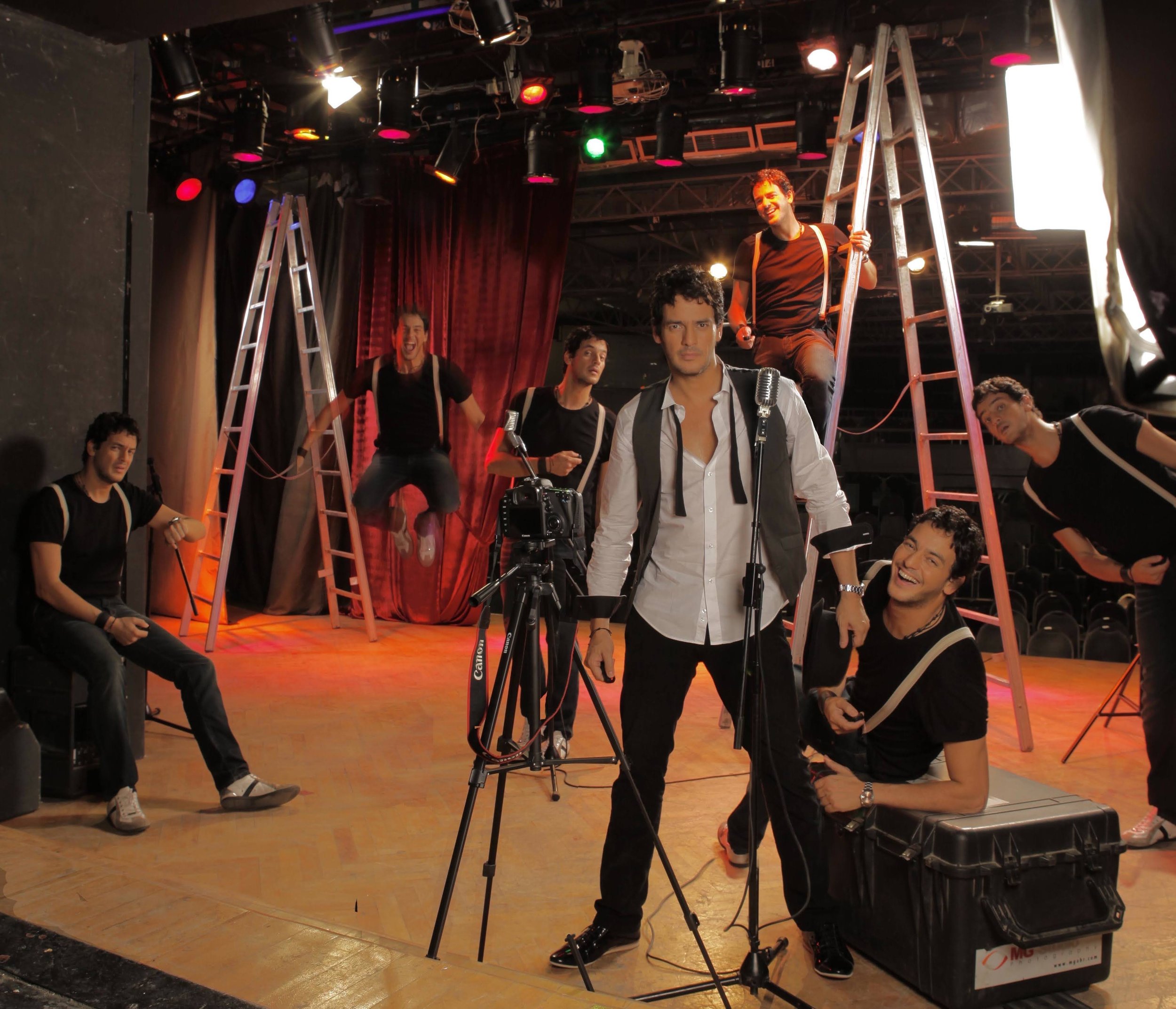 A group of performers on stage during a theatrical production, with some holding ladders, surrounded by stage equipment and lighting, and one man in the foreground looking serious with microphones in front of him.
