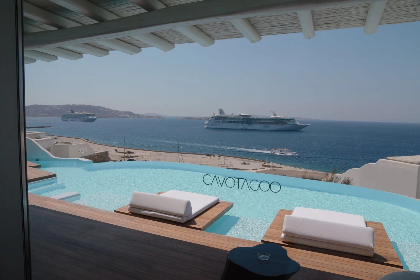View from a balcony overlooking a curved infinity pool with white towels on wooden lounges, sea with two cruise ships, and clear sky in the background.
