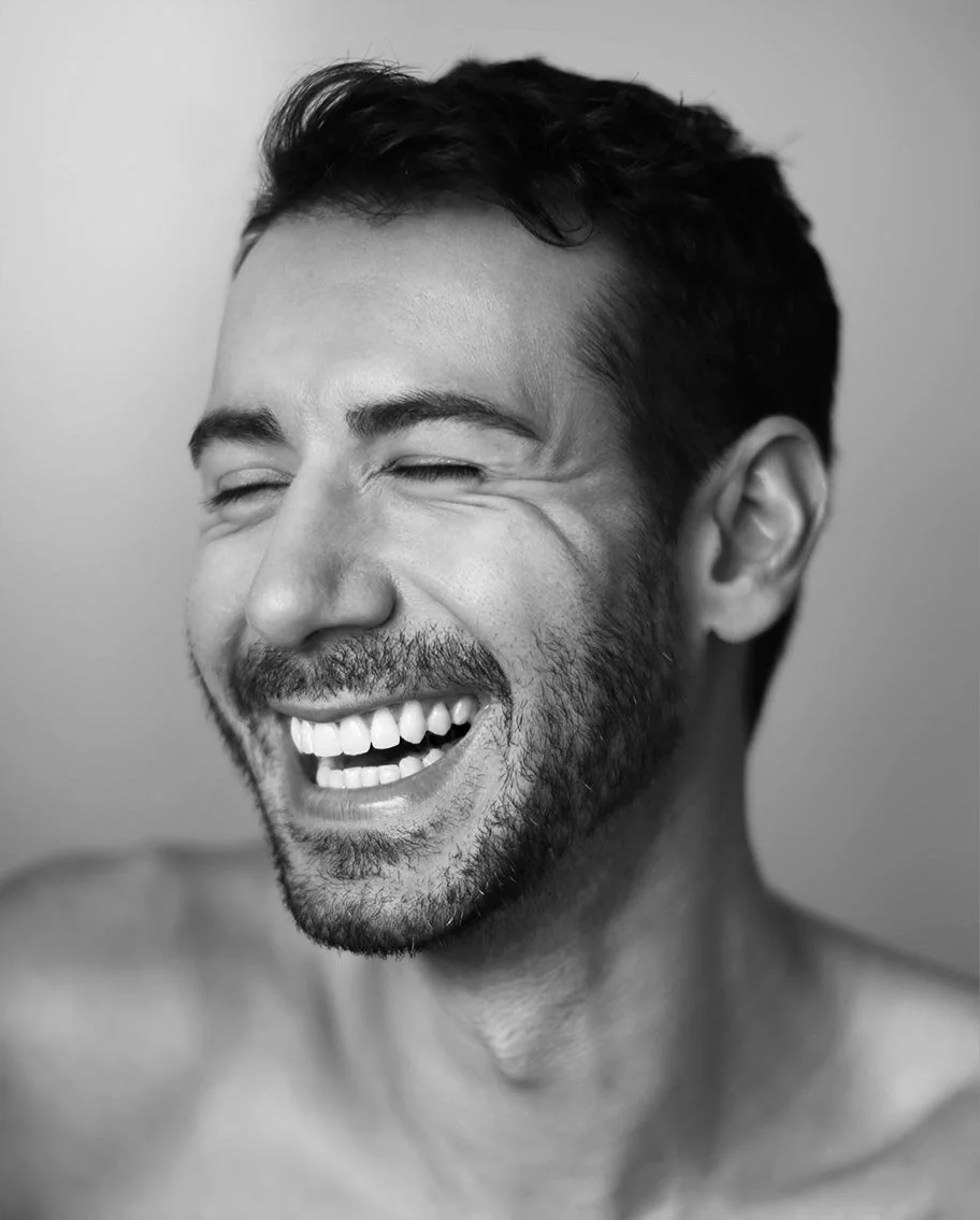 A black and white close-up photo of a smiling man with closed eyes, short dark hair, and a beard.