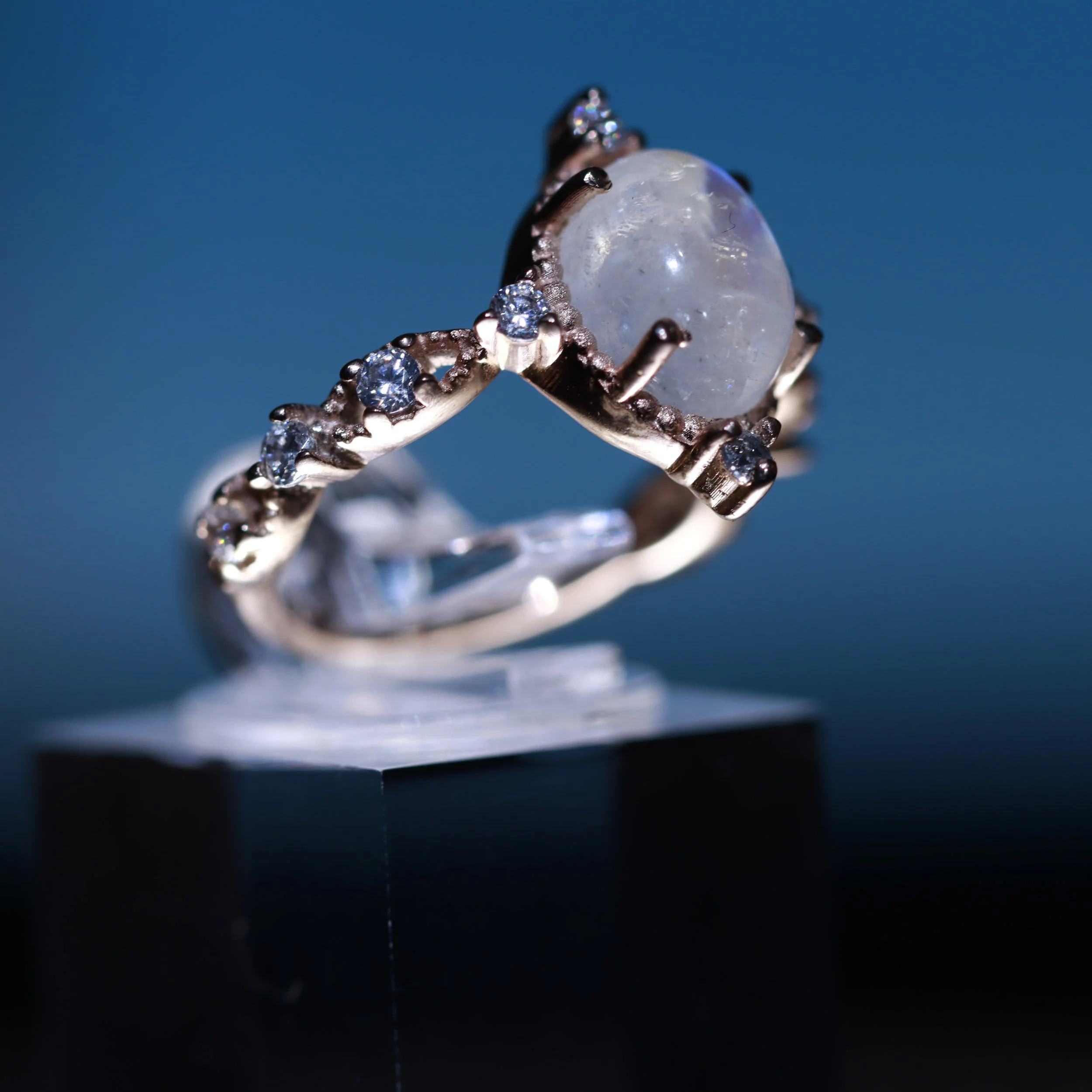 Close-up of a silver ring with a large central moonstone surrounded by smaller diamonds, set against a blue background.