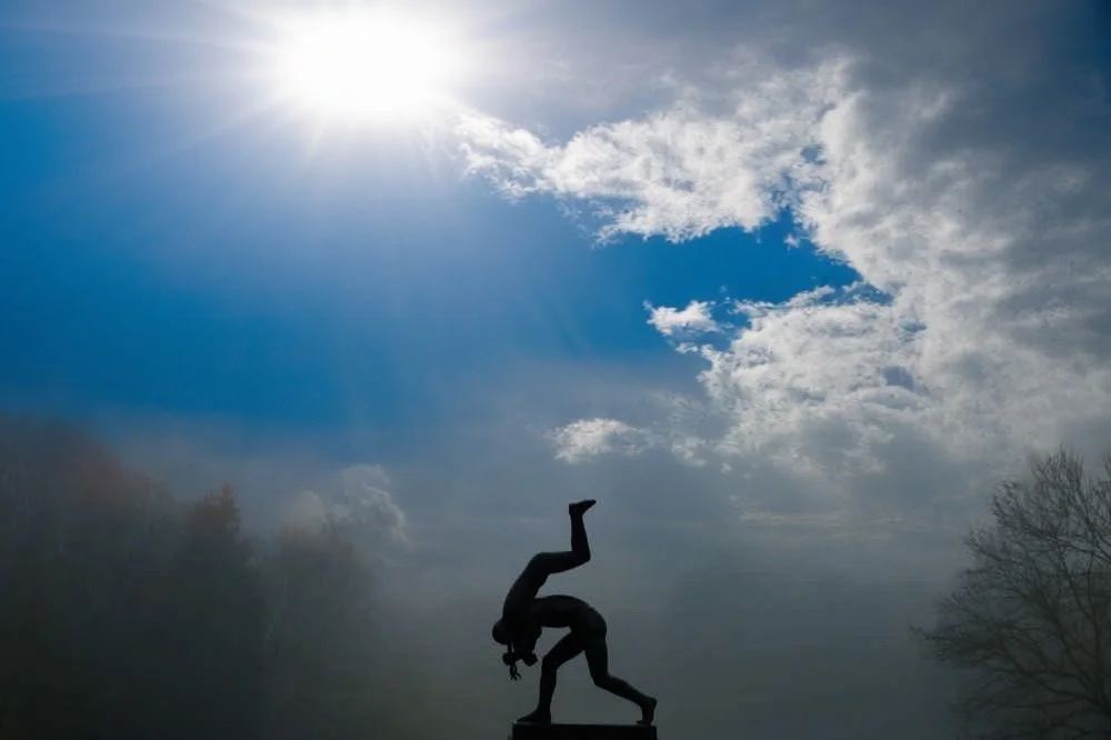 Silhouette of a person performing a headstand on a platform outdoors, with a cloudy sky and sun shining overhead.
