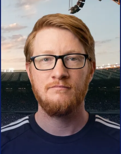 A man with red hair, glasses, and a red beard standing outdoors in front of a stadium, wearing a dark sports jersey.
