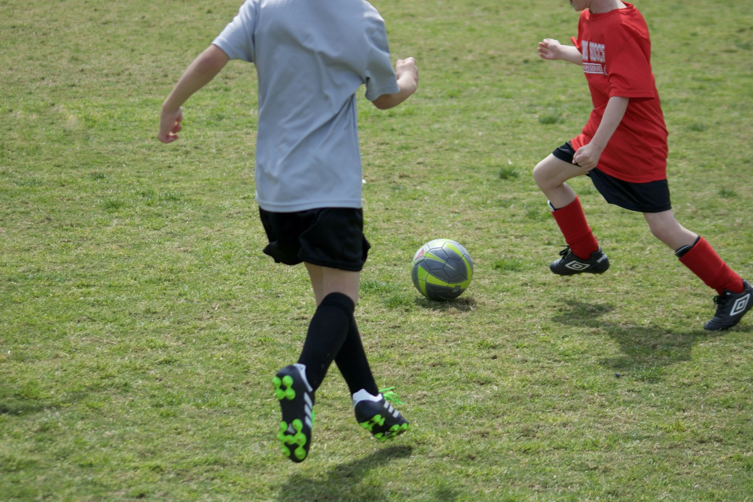 Two children playing soccer on a grassy field, one wearing a gray shirt and black shorts, the other in a red shirt and black shorts, with a green and gray soccer ball between them.