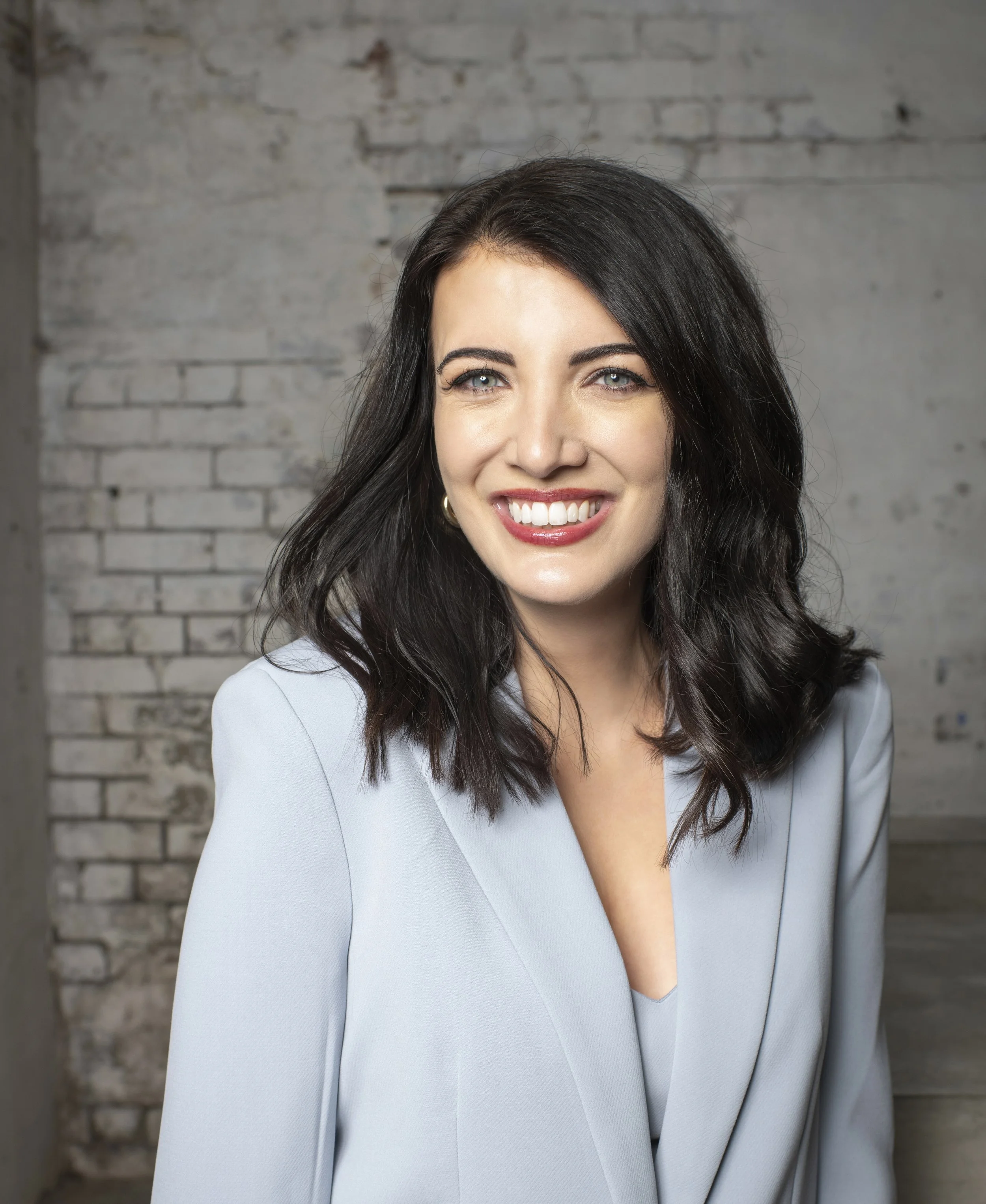 A woman with dark hair, light blue eyes, and a bright smile, wearing a light blue blazer, standing against a gray brick wall background.