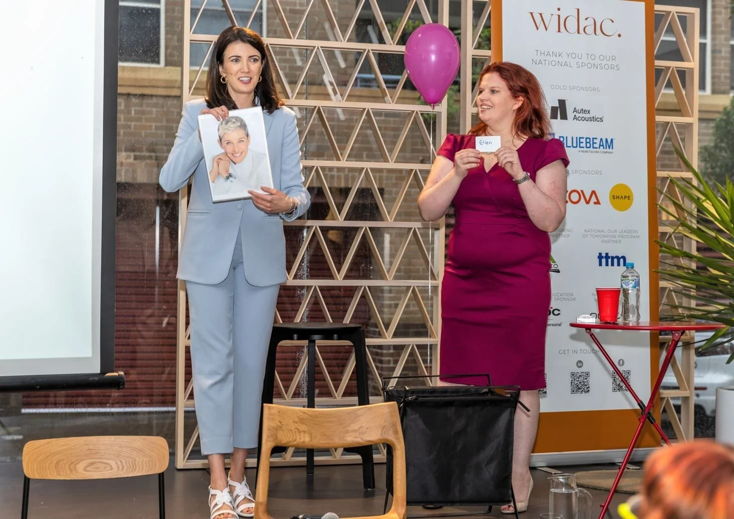 Two women are standing indoors in front of a large sponsor banner, engaging in a presentation or award event. The woman on the left is holding a magazine or photo of a smiling woman with short gray hair, dressed in white. The woman on the right is holding a card with the name "Ellen" written on it. A small round table with a water bottle and red cup, and a chair, are visible nearby, with balloons and a decorative wooden lattice in the background.