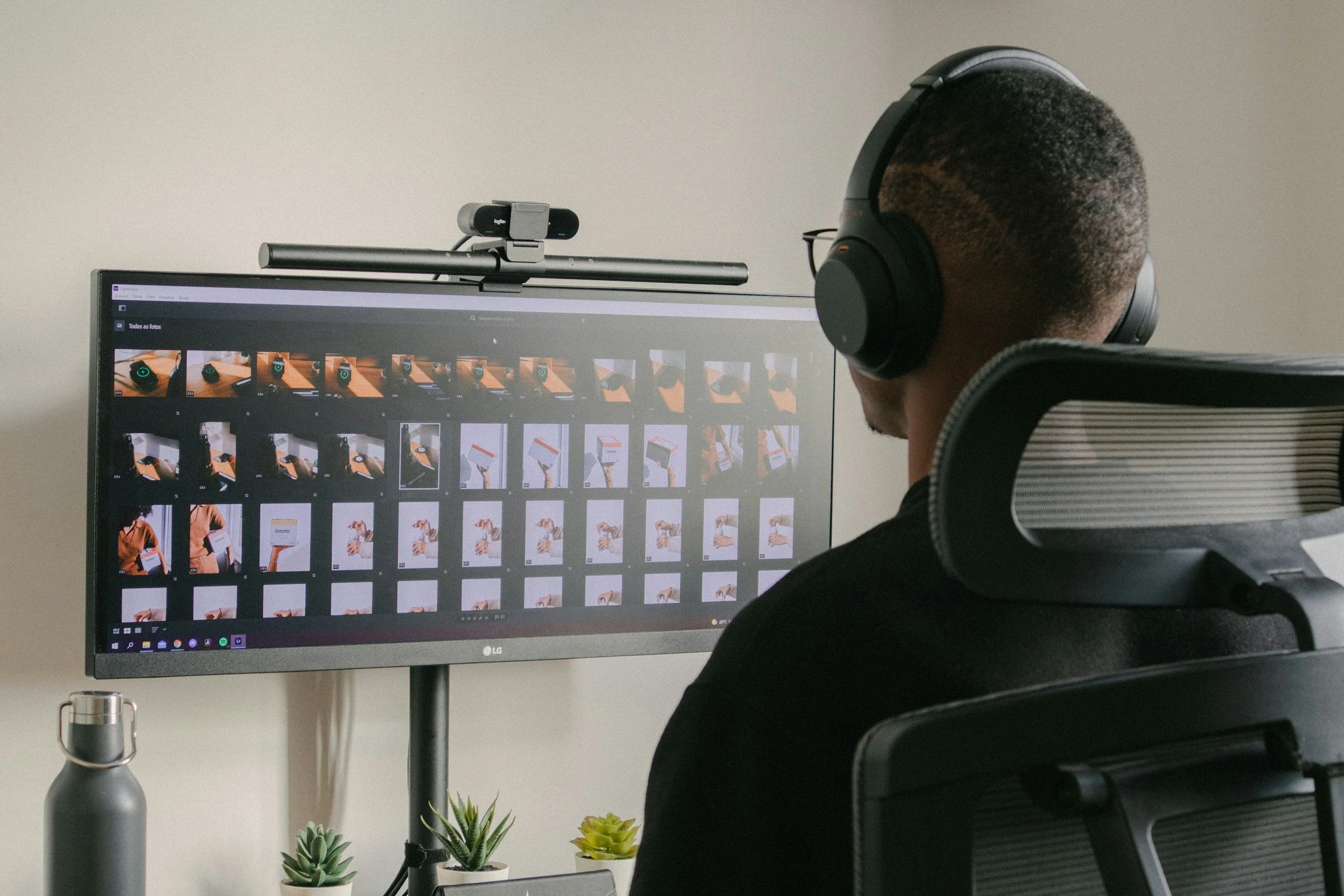 A person with short, curly hair and glasses wearing over-ear headphones sits in front of a computer monitor displaying a photo gallery. The monitor shows multiple rows of photographs, possibly of a product or object, with a search bar at the top. There is a webcam mounted on top of the monitor. On the desk, there are several potted succulent plants and a gray water bottle.