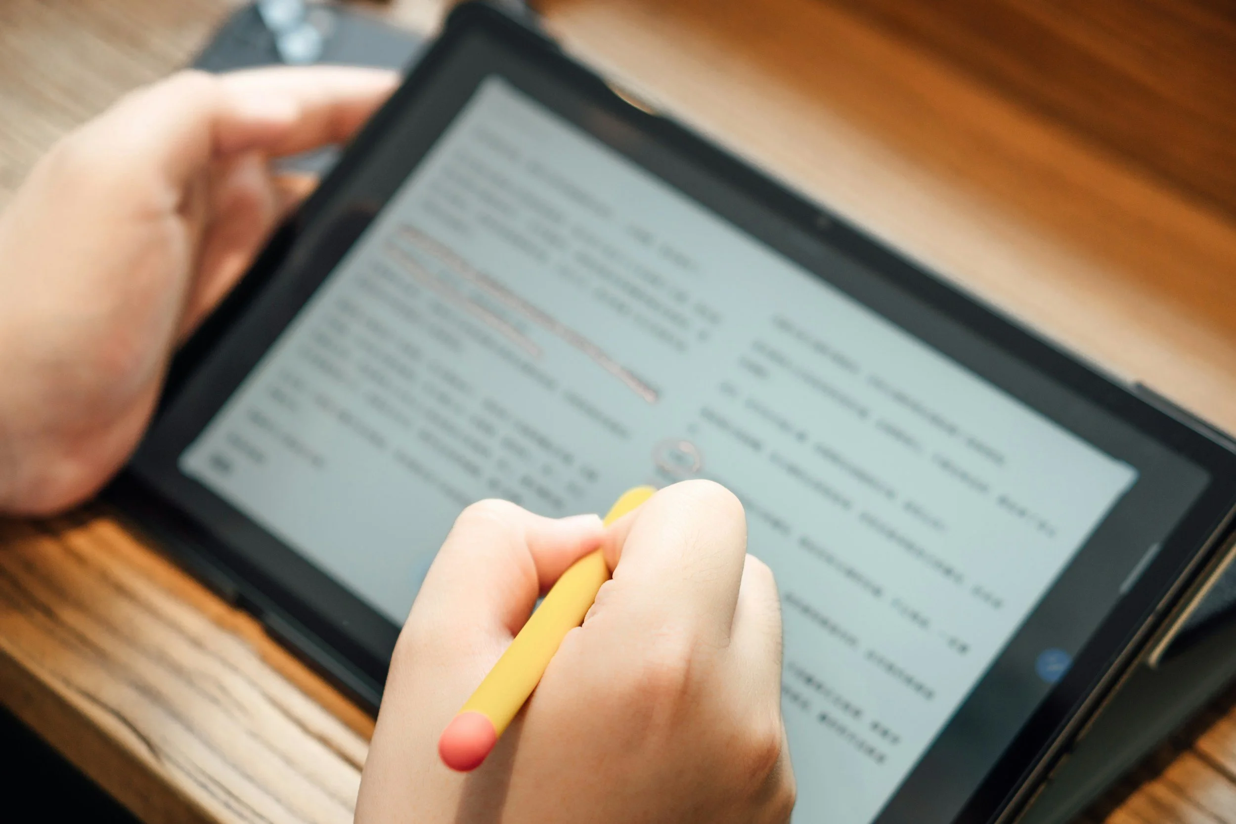 A person's hand holding a yellow stylus and writing on a digital tablet with a stylus, which is placed on a wooden surface. The screen displays text that appears to be an email or document.
